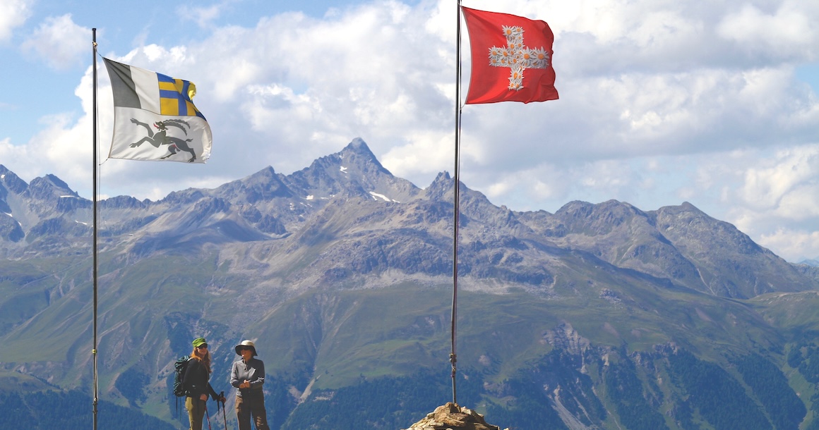 Two hikers between two flags with peaks in the background.