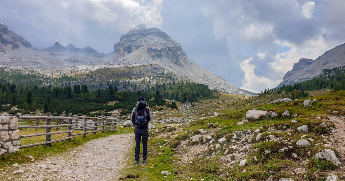 A man with a big hiking backpack hiking along a gravelled road in Italian Dolomites.